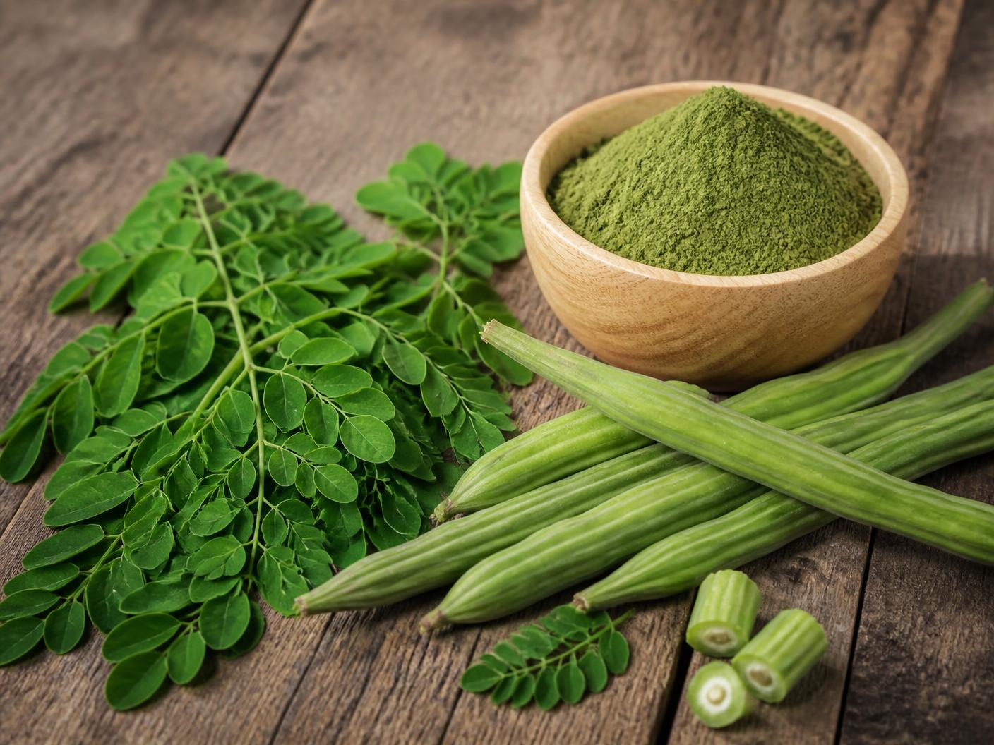 Fresh moringa leaves and drumstick pods beside a bowl of moringa powder